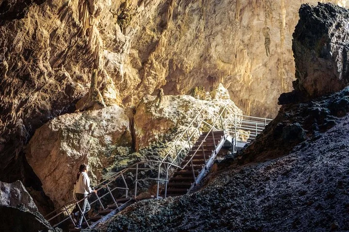 Eine Frau steigt eine Metalltreppe in einer riesigen, beleuchteten Höhle mit Stalaktiten hinauf.